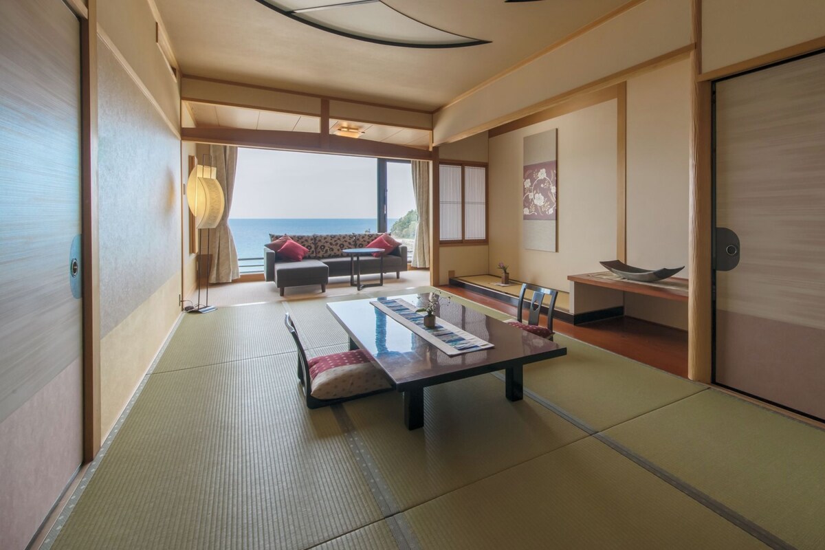 Una habitación tradicional japonesa con suelo de tatami, una mesa baja con una bandeja, un sofá con cojines y vistas al mar a través de amplias ventanas.