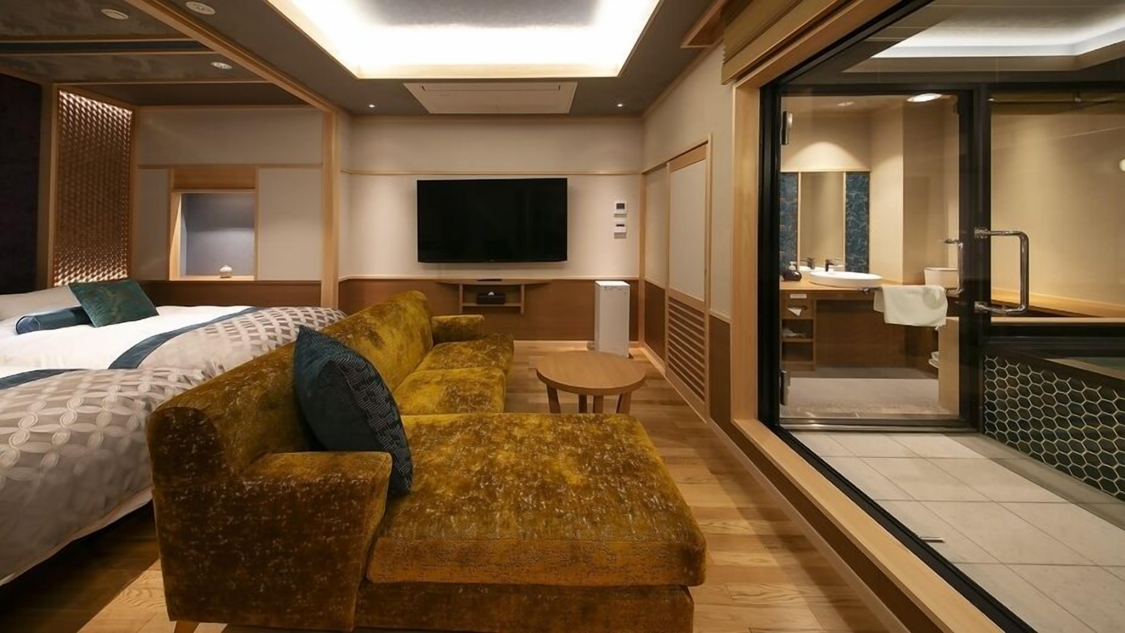 Une chambre d’hôtel comprenant un lit, un canapé, une télévision, une petite table et une salle de bain visible à travers une porte en verre.