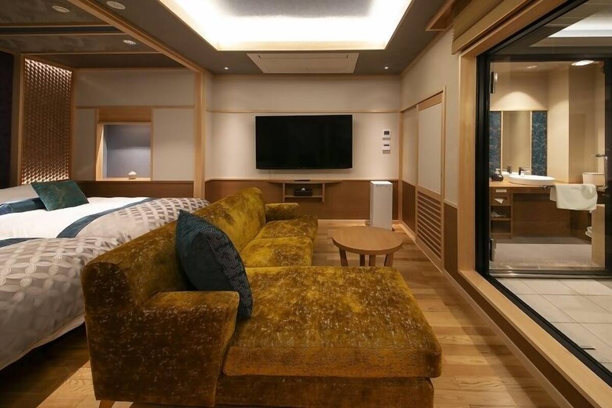 A hotel room with a bed, a sofa, a TV, a small table, and a bathroom visible through a glass door.