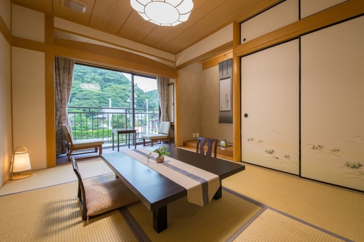 Ein traditioneller japanischer Raum mit Tatami-Böden, einem niedrigen Tisch mit Stühlen, einer Schiebetür mit Kunstwerk und Blick auf Grünfläche.