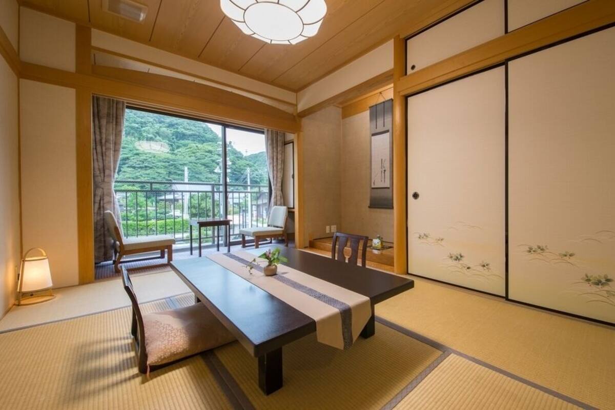 A traditional Japanese room with tatami flooring, a low table with chairs, a sliding door with artwork, and a view of greenery outside.