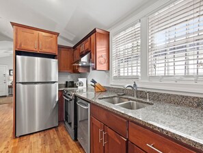 A modern kitchen with stainless steel appliances, wooden cabinets, granite countertops, and a large window with blinds.
