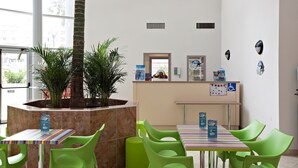 Un espacio interior moderno con mostrador de recepción, sillas verdes, una mesa a rayas y una gran planta en maceta.