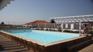Una piscina rectangular con una terraza de madera alrededor, un área de estar cubierta y un edificio con techo de tejas rojas al fondo.