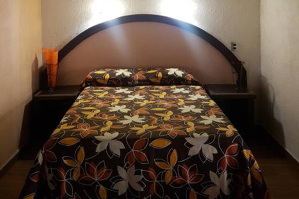 Une chambre à coucher avec une tête de lit en bois, un lit recouvert d’une couvre-lit à motifs floraux, deux lampes de chevet et une croix accrochée au mur.