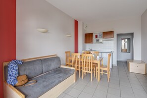A modern living room with a grey sofa, wooden dining table, and chairs, a kitchenette in the background, and a red accent wall.