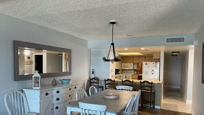 Un comedor con mesa de madera y sillas blancas, una cocina con armarios de madera y un refrigerador, y un techo con textura.