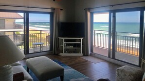 Una habitación con ventilador de techo, televisor en un soporte y un amplio ventanal corredero que da a un balcón con vista a la playa.