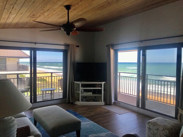 A room with a ceiling fan, a TV on a stand, and a large sliding glass door leading to a balcony with a view of the beach.