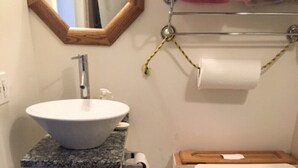 A bathroom with a white sink, a toilet, a blue trash bin, and a wooden shelf above the toilet.