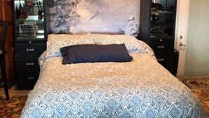 A bedroom with a patterned bedspread, a dark pillow, a headboard with a winter scene, and a nightstand with a lamp and a vase of flowers.