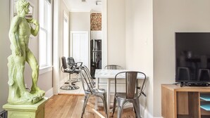 Uma área de refeições com mesa e cadeiras, uma estátua, uma televisão e uma cozinha ao fundo.