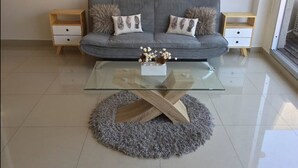 Ett modernt vardagsrum med en grå soffa, två sidobord, ett soffbord i glas och en grå, lurvig matta.
