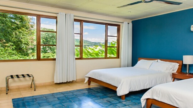 A hotel room with two beds, a window with a view of greenery, a blue tiled floor, and an air conditioning unit on the wall. - Montebrisa Boutique B&B (Matagalpa)