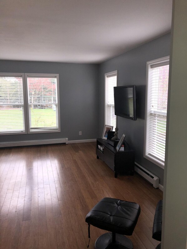 Una habitación amplia con piso de madera, un reposapiés negro, un televisor de pantalla plana en la pared y dos ventanas con persianas.