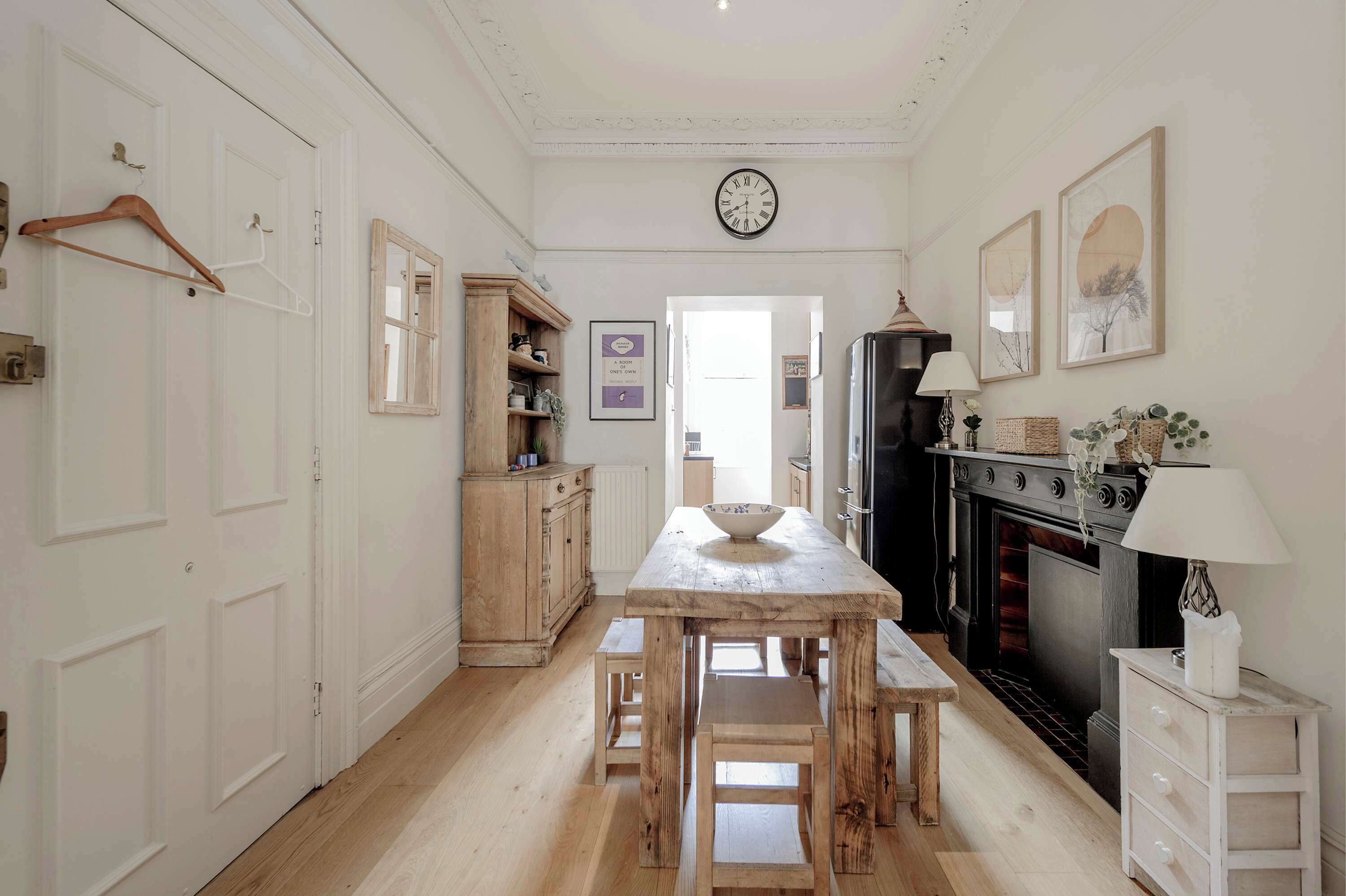 Una cocina con una mesa de madera, bancos, un perchero de madera colgante, un reloj, una chimenea y un armario blanco.