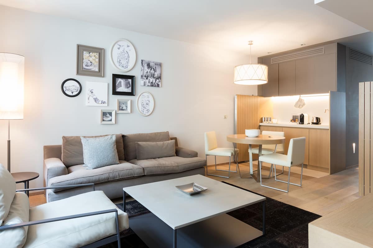 Uma sala de estar moderna com um sofá, cadeiras, uma mesa de centro e um espaço de jantar com mesa e cadeiras. Uma cozinha é visível ao fundo.