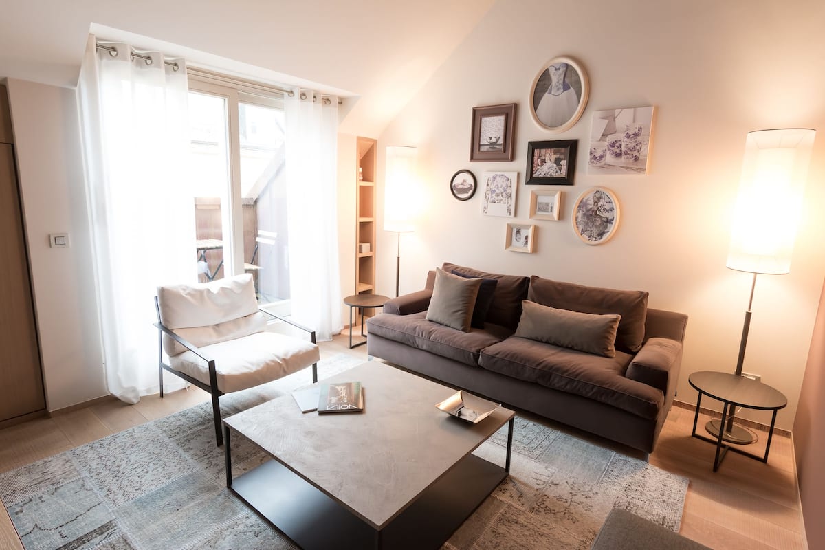 Uma sala de estar moderna com sofá, mesa de centro e luminária. Há quadros nas paredes e uma janela com cortinas.