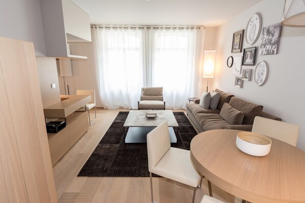 A modern living room with a sofa, a coffee table, and a dining table. There is a large window with curtains, a wall-mounted shelf, and several framed pictures. - Guillaume Suites (Luxembourg City)