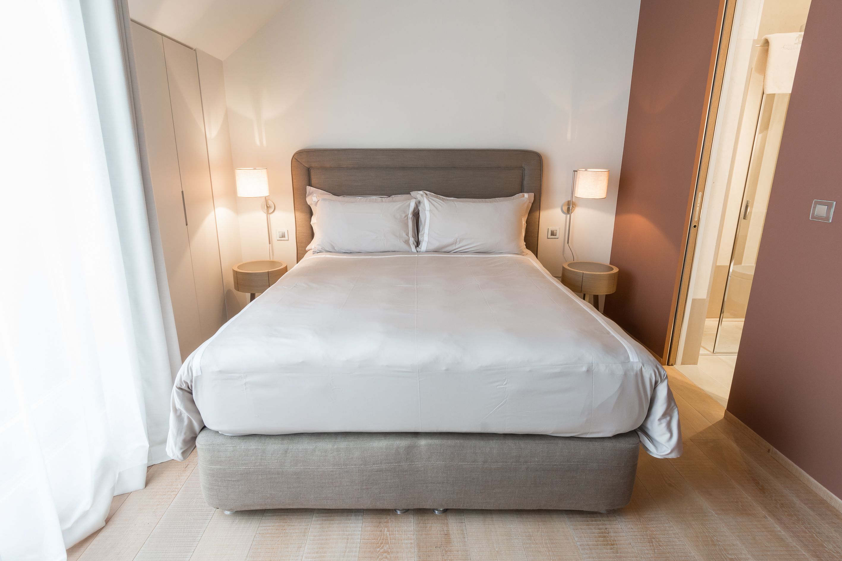 Ein ordentlich bezogenes Bett mit grauem Kopfteil und weißen Laken, flankiert von zwei Nachttischlampen auf passenden Beistelltischen.