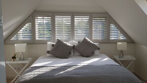 Ein Schlafzimmer mit schrägen Wänden, einem Bett mit Strickdecke, zwei Nachttischen mit Lampen und einem Fenster mit Jalousien.