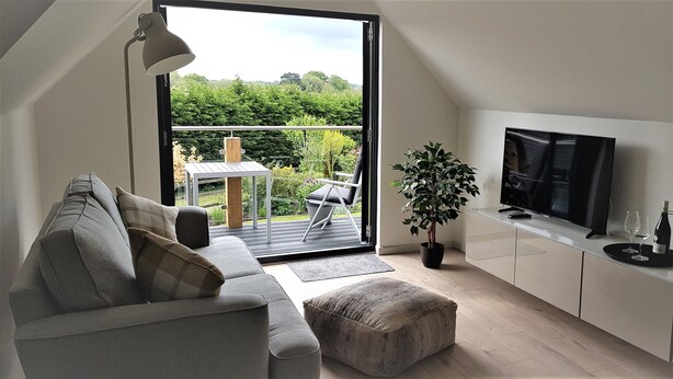 Ein modernes Wohnzimmer mit Sofa, Hocker, Fernseher und einem Balkon mit Tisch und Stühlen.