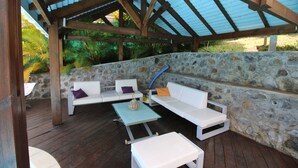 Une terrasse extérieure avec des murs en pierre, des meubles en bois et un espace abrité doté d’un toit en métal.