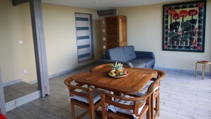Un coin repas avec une table et des chaises en bois, un panier de fruits, un canapé gris et une peinture murale aux couleurs vives.