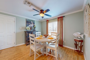 A dining area with a round table, white chairs, a ceiling fan, and a window with curtains.