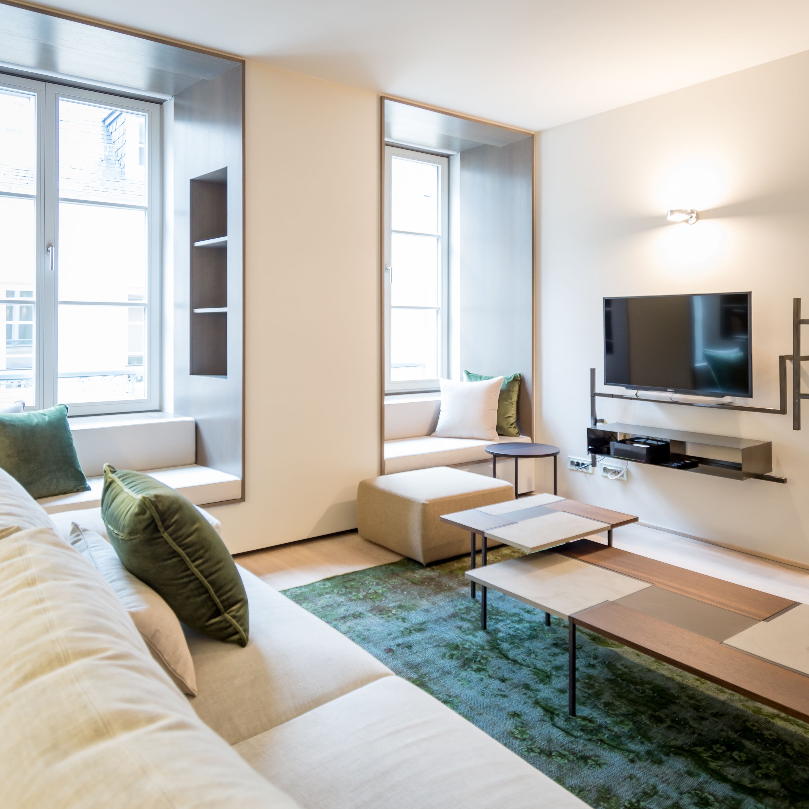 A modern living room with a sofa, a coffee table, a TV mounted on the wall, and large windows.