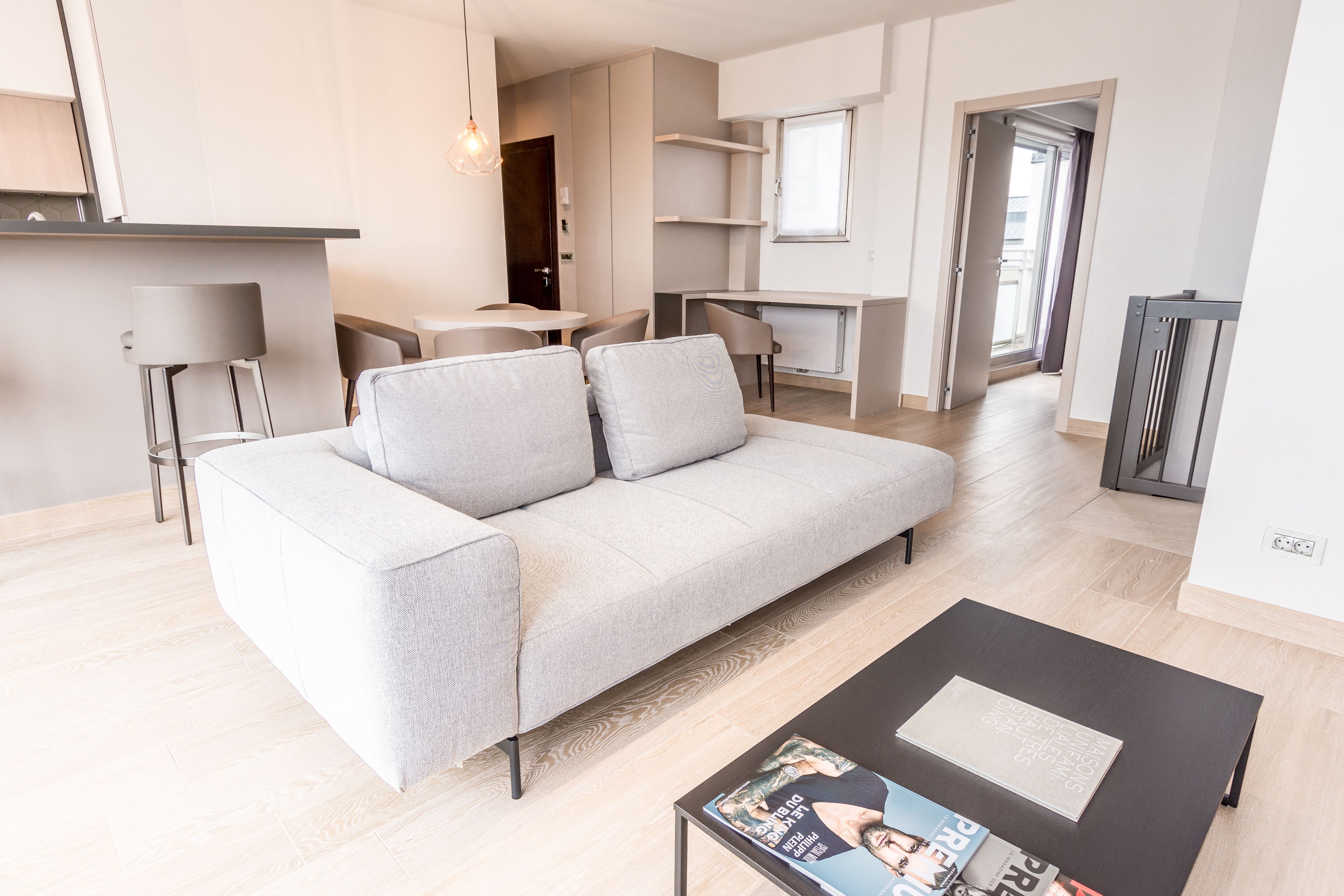 A modern living room with a grey sofa, a coffee table with magazines, and a kitchen area in the background.