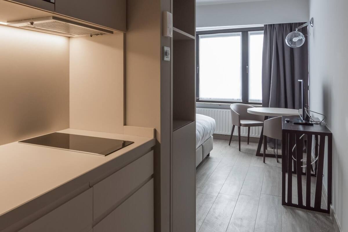 Een moderne hotelkamer met kitchenette, een bed, een bureau met stoel en een raam met gordijnen.
