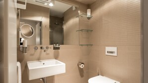 Une salle de bain moderne avec un lavabo, des toilettes et une douche, le tout de couleur blanche. On trouve des étagères en verre au-dessus de la douche et un miroir au-dessus du lavabo.