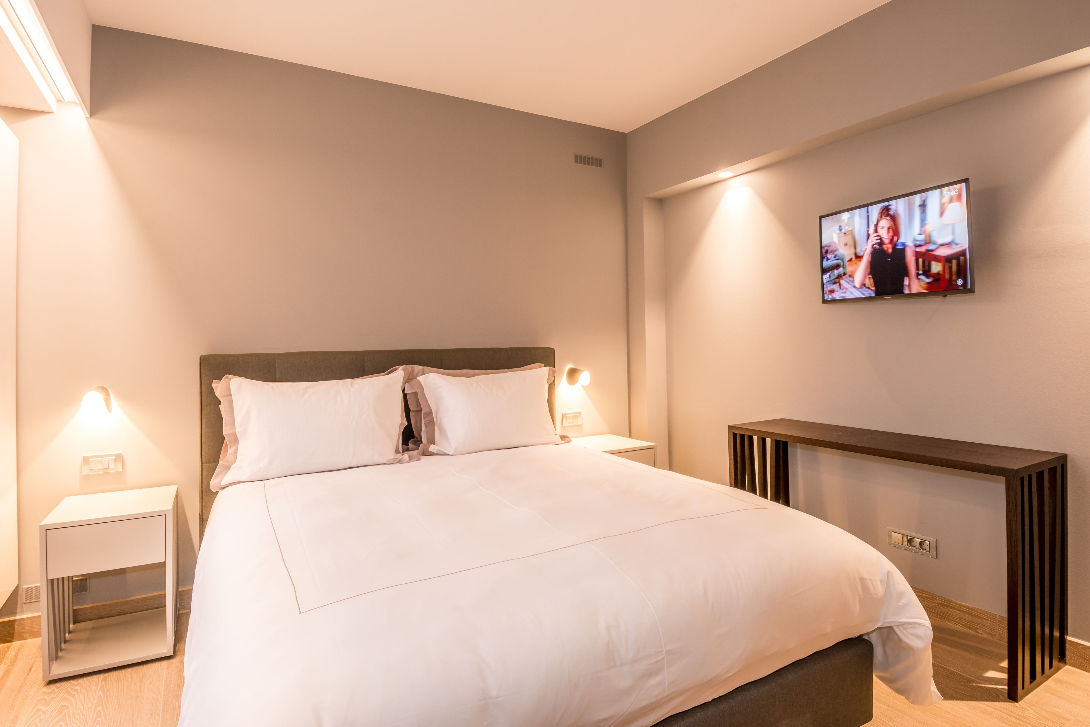 A modern hotel room with a large bed, bedside lamps, a TV mounted on the wall, and a wooden console table.