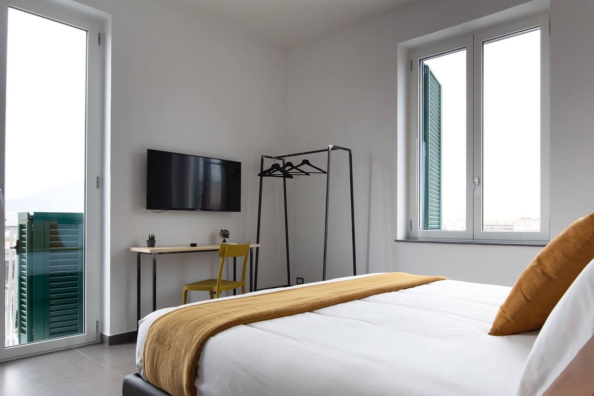 A bedroom with a bed, a desk, a chair, a TV, and a window.