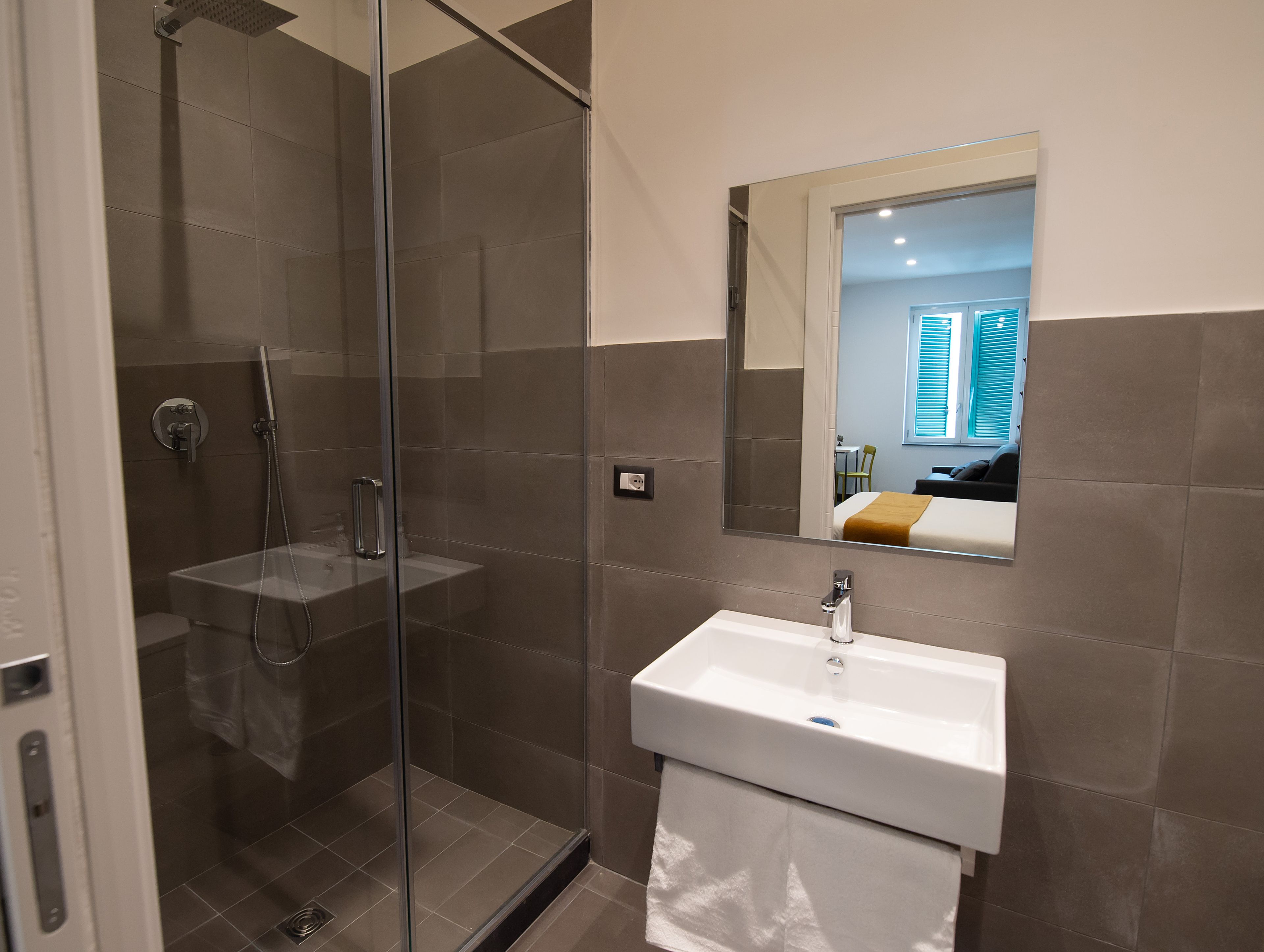Een moderne badkamer met een glazen douchecabine, een wit wastafelmeubel en een spiegel die een slaapkamer met een bed en raam weerspiegelt.