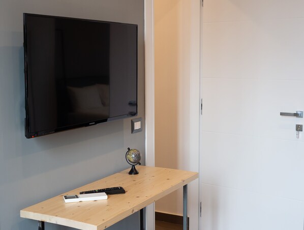 A room with a mounted flat-screen TV, a wooden desk with a globe and remote controls, and a white door. - GB Vesuvius Napoli (Naples)