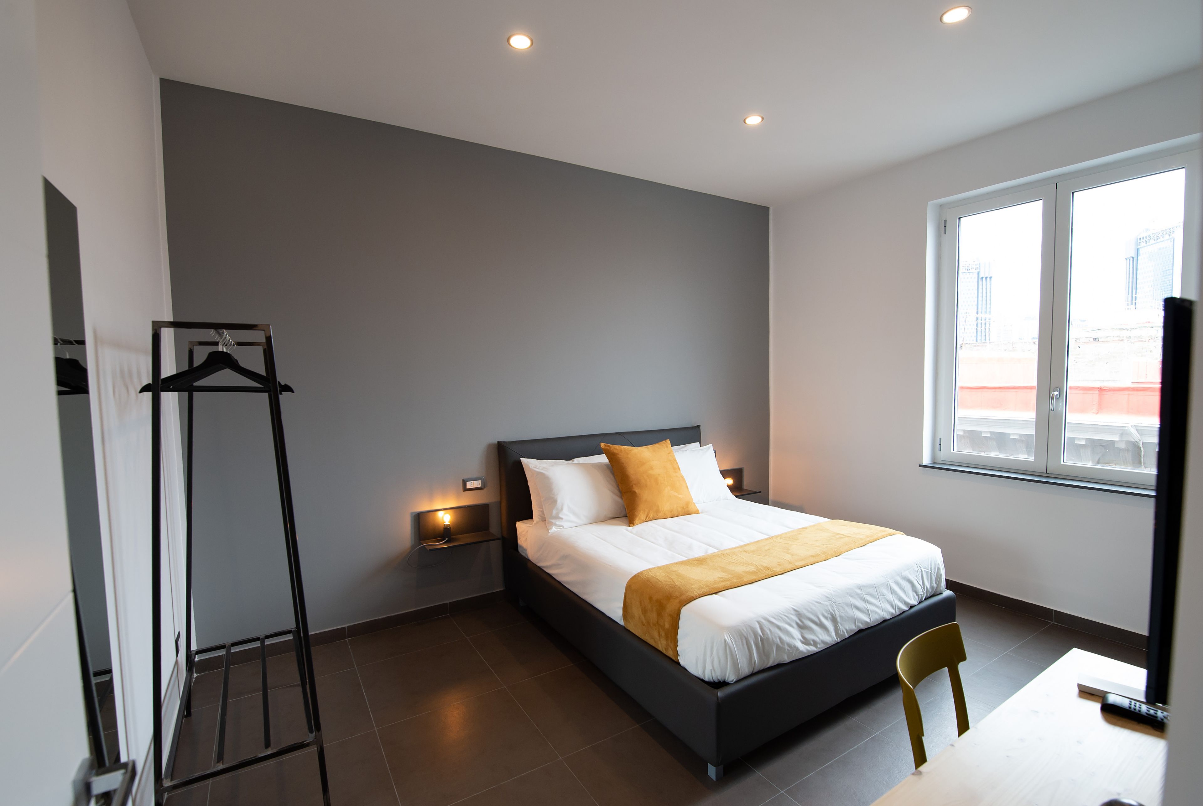 Ein modernes Hotelzimmer mit einem großen Bett, einem Nachttisch, einem Sessel, einem Kleiderständer und einem Fenster mit Blick auf die Stadt.