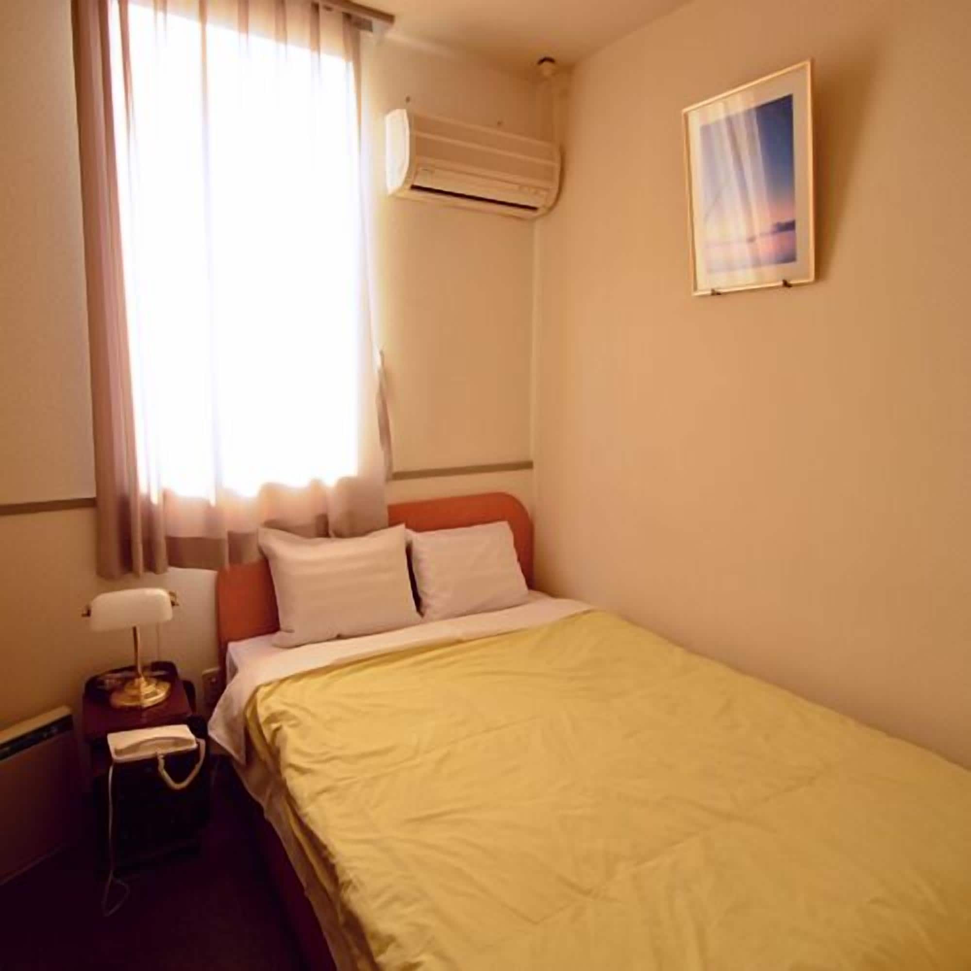 A hotel room with a bed, a bedside table with a lamp, a telephone, and a window with curtains.