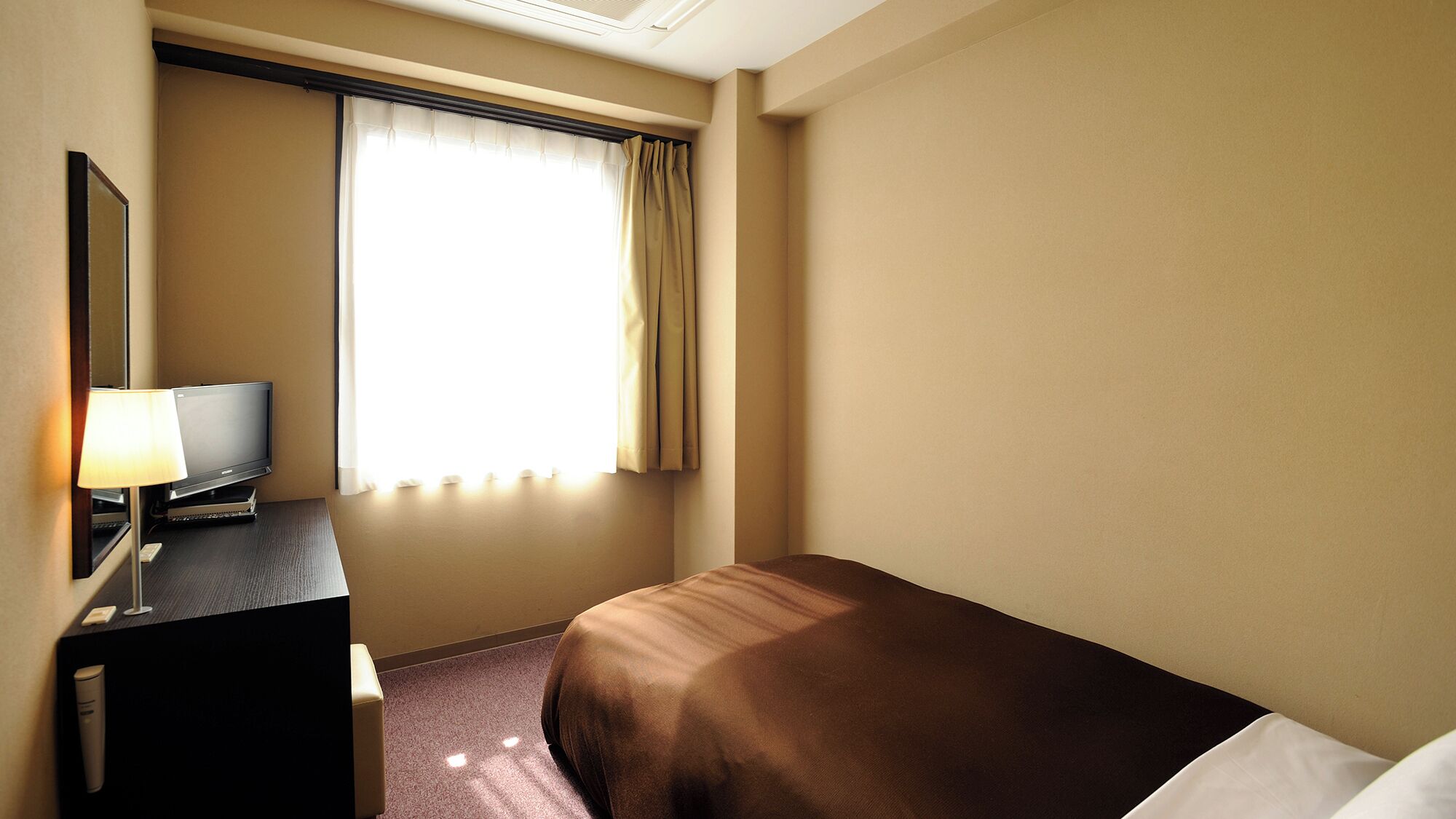 A hotel room with a bed, a desk with a lamp, a television, and a window with curtains.