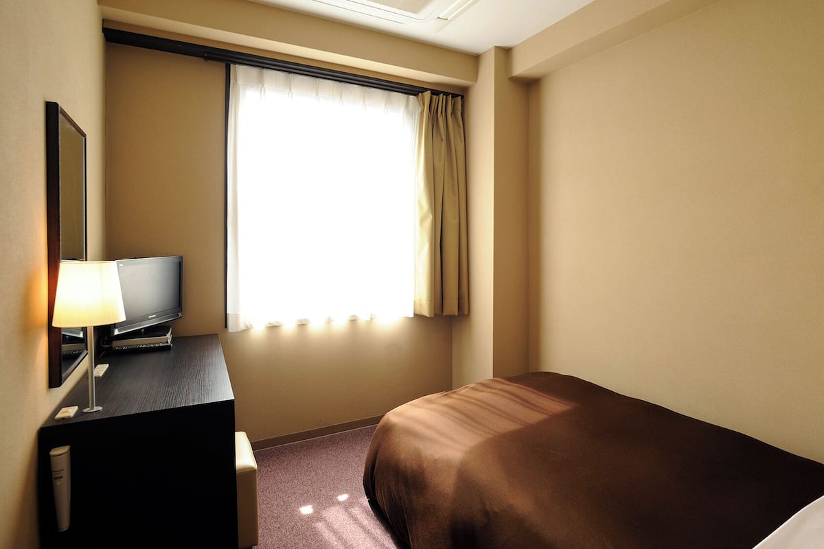 Une chambre d’hôtel avec un lit, un bureau avec une lampe, une télévision et une fenêtre avec des rideaux.