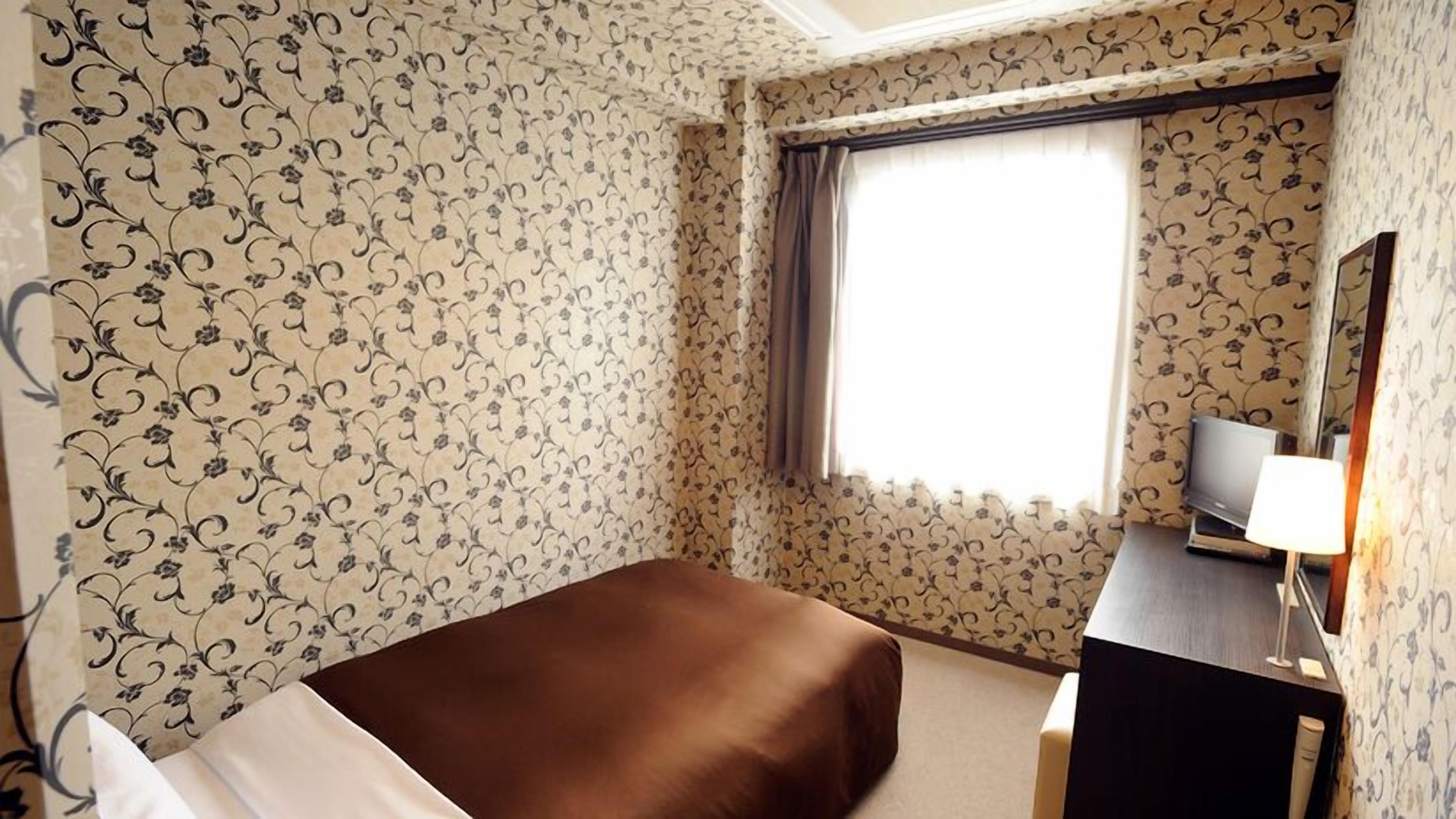 Habitación de hotel con papel tapiz floral, una cama, un escritorio con lámpara y un televisor.