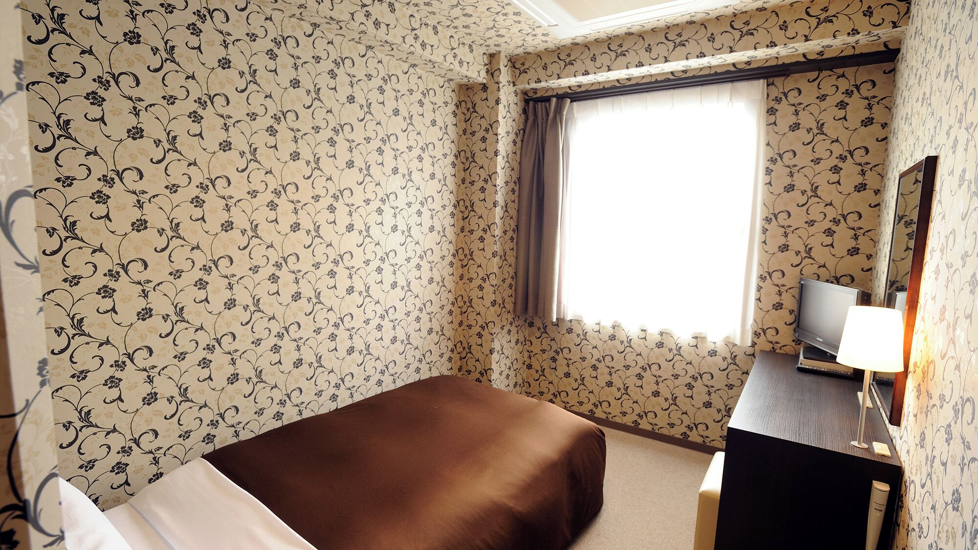 A hotel room with floral wallpaper, a bed, a desk with a television, and a window with curtains.