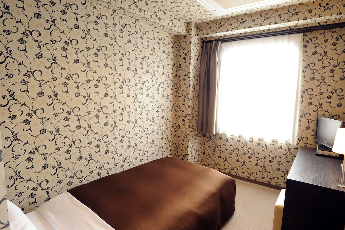 Une chambre d’hôtel avec un papier peint à motifs floraux, un lit, un bureau avec une télévision et une fenêtre avec des rideaux.