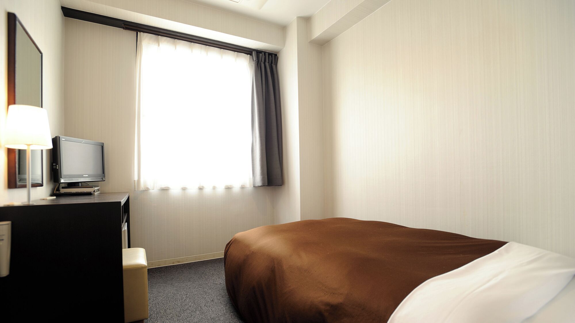 A hotel room with a bed, a desk with a television, a lamp, and a window with curtains.