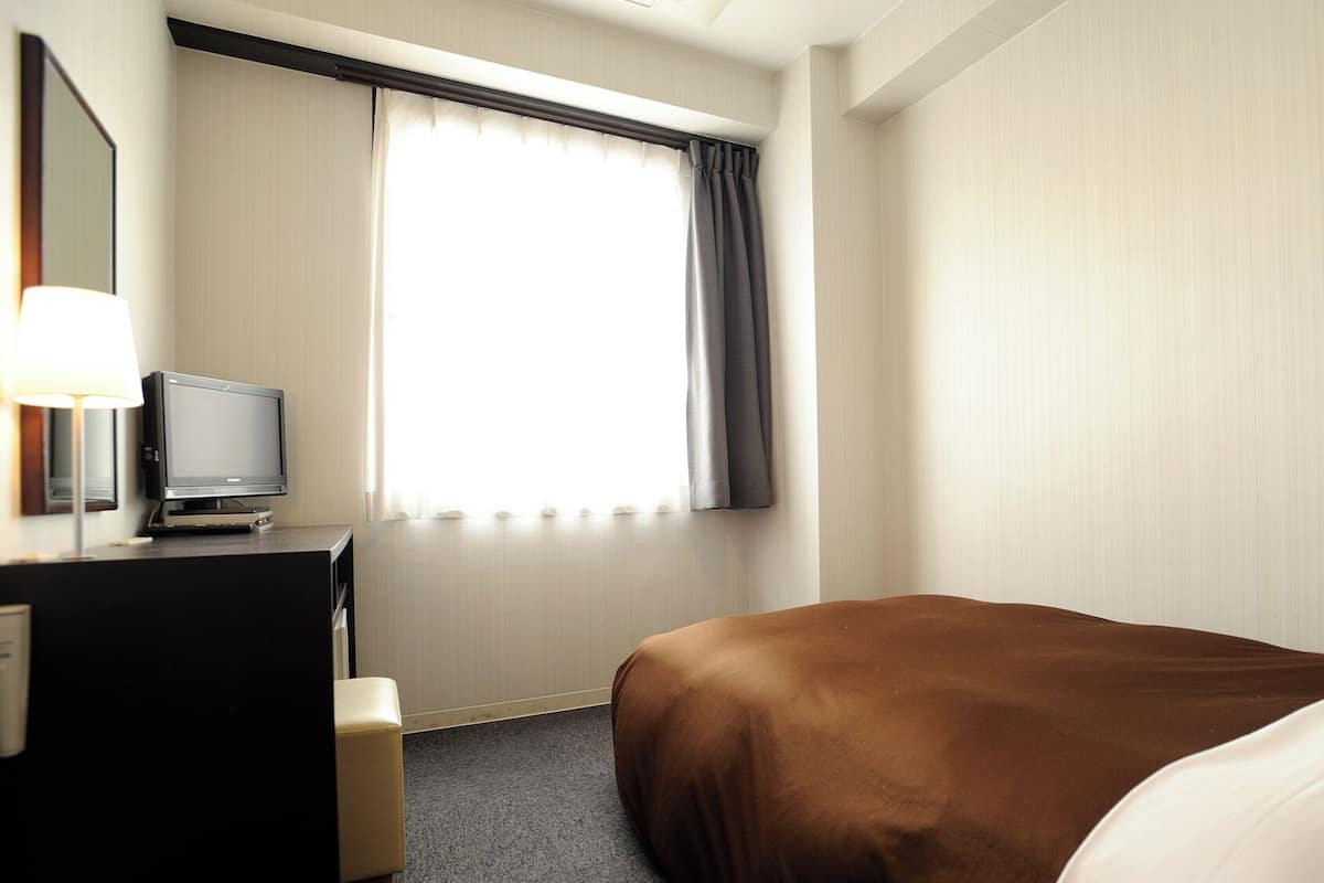 Une chambre d’hôtel avec un lit, un bureau avec une télévision, une lampe et une fenêtre avec des rideaux.