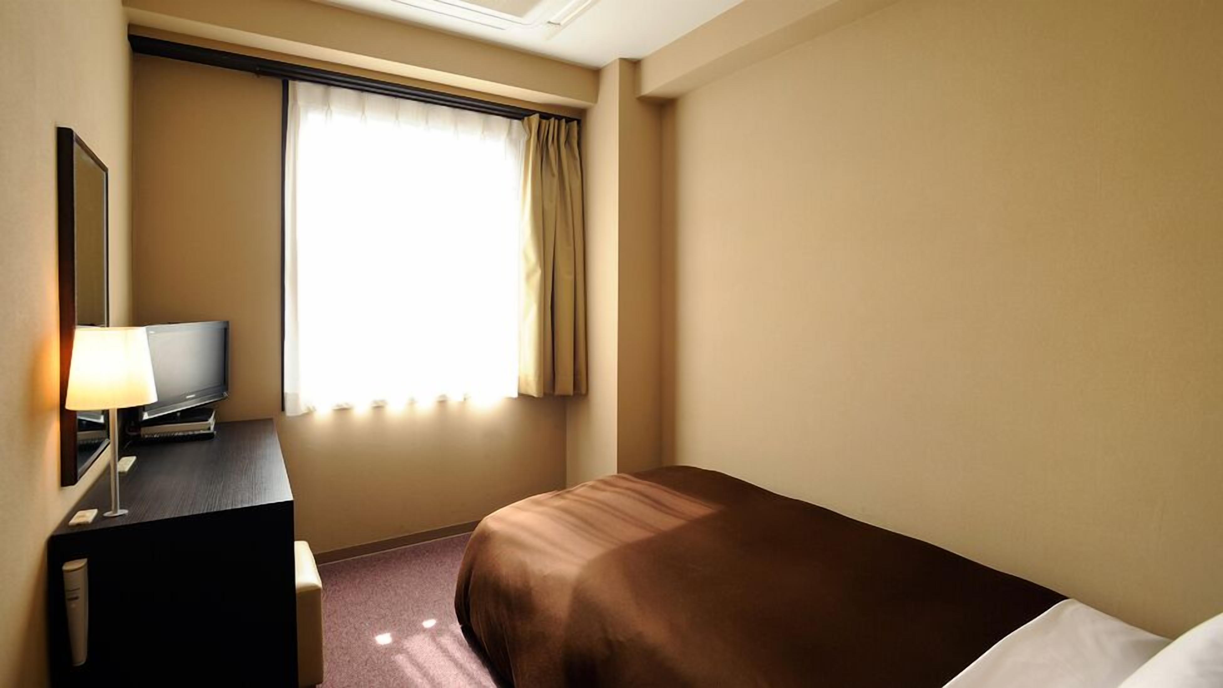 Una habitación de hotel con cama, escritorio con lámpara, televisión y ventana con cortinas.