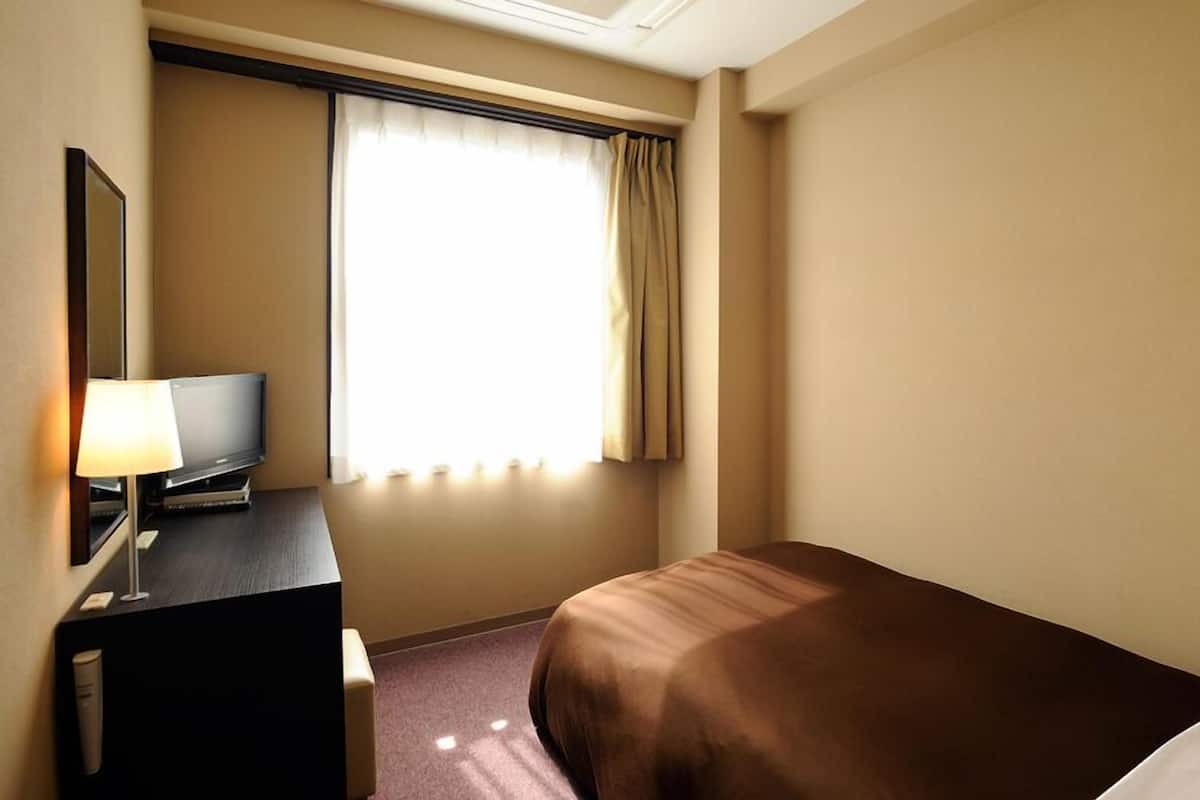 Une chambre d’hôtel avec un lit, un bureau avec une lampe, une télévision et une fenêtre avec des rideaux.
