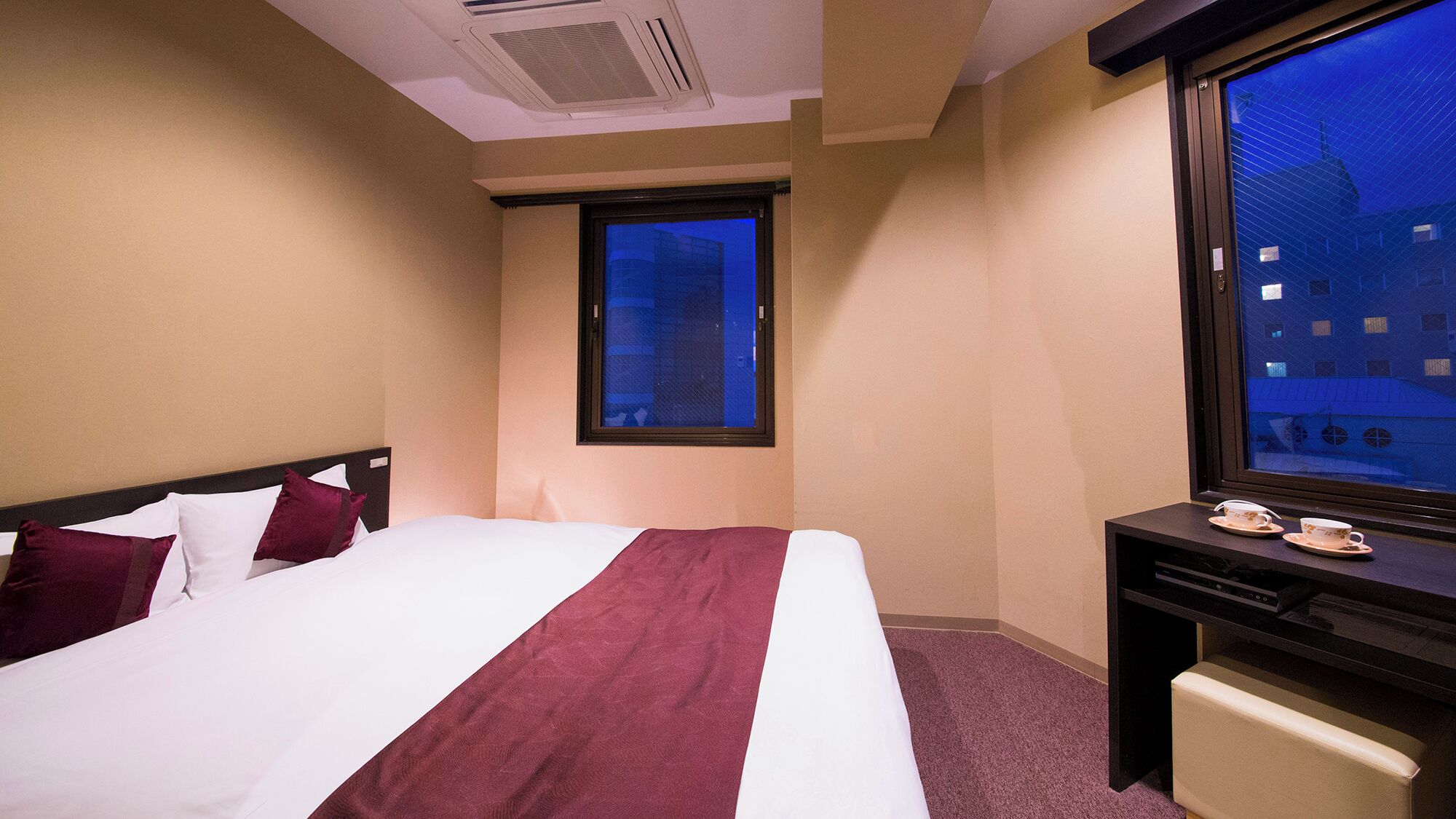 A hotel room with a bed, a window with a view of buildings, a desk with a computer, and a small table with cups.
