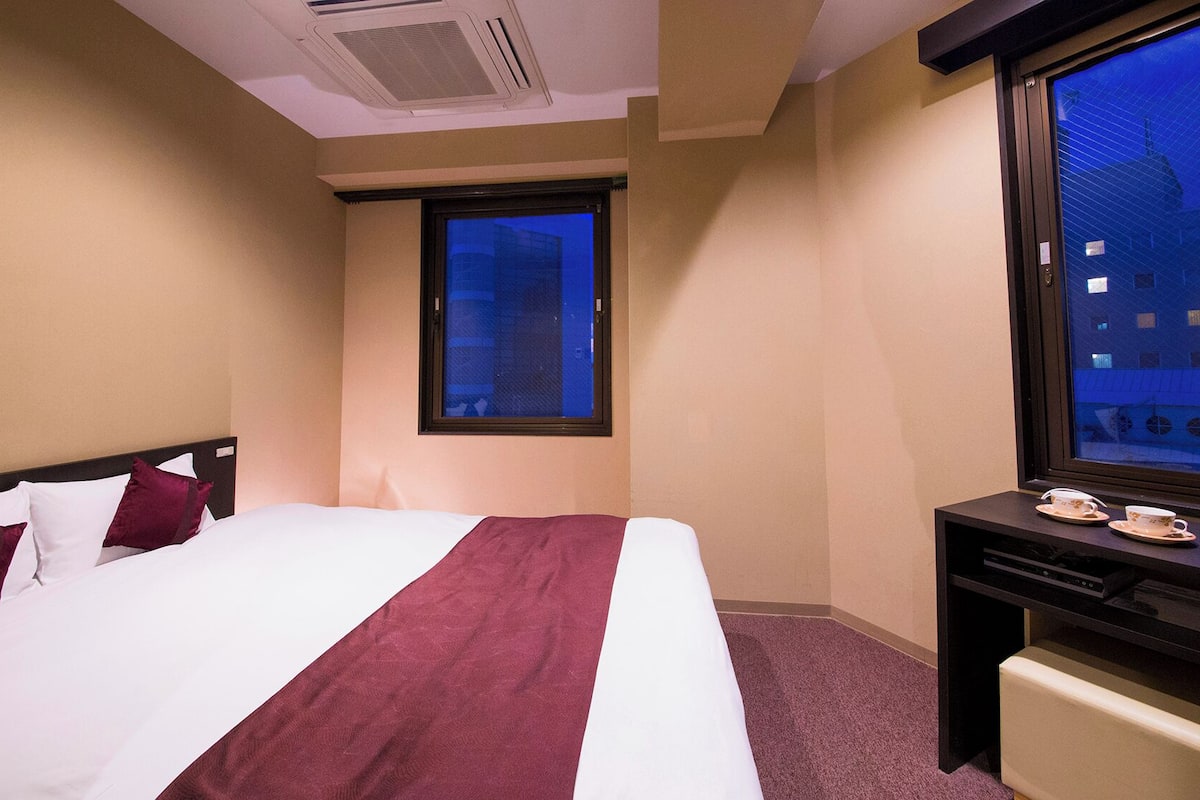 Une chambre d’hôtel avec un lit, une fenêtre donnant sur des immeubles, un bureau avec un ordinateur et une petite table avec des tasses.
